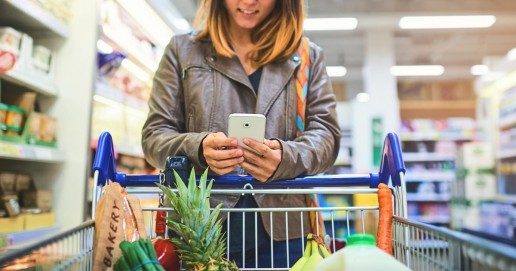 Eine Frau im Supermarkt schiebt ihren Einkaufswagen und schaut auf ihr Handy